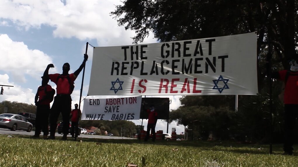 An image of individuals associated with the National Socialist Front holding a sign that says "The Great Replacement Is Real," in black and red lettering and two Stars of David. The "Is Real" is also meant to be read as "Israel." There is another sign in the background that says "End Abortion, Save White Babies" in black and red text.