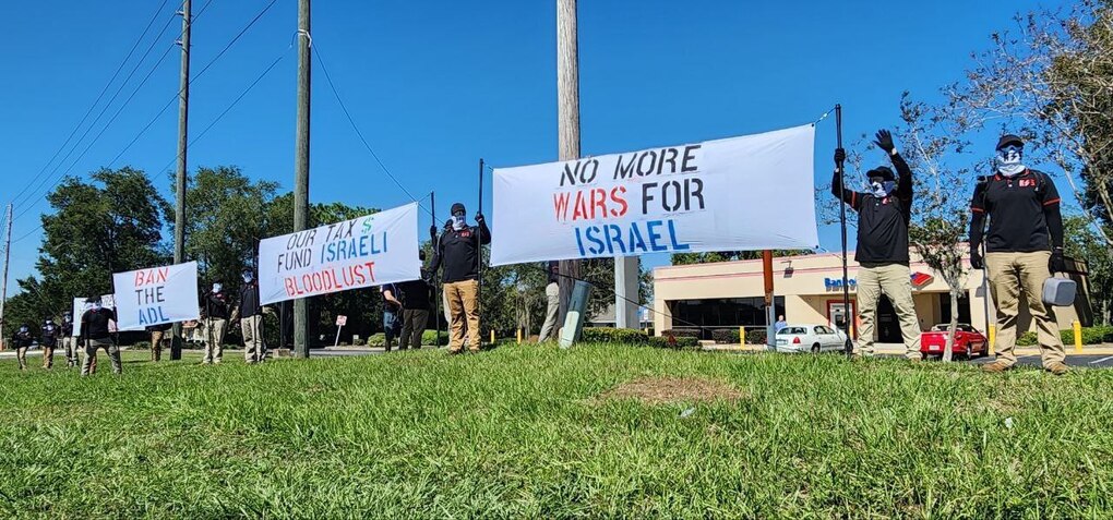 An image of individuals associated with the National Socialist Front holding signs that say "Ban the ADL," "Our Tax $ Fund Israeli Bloodlust" and "No More Wars for Israel."